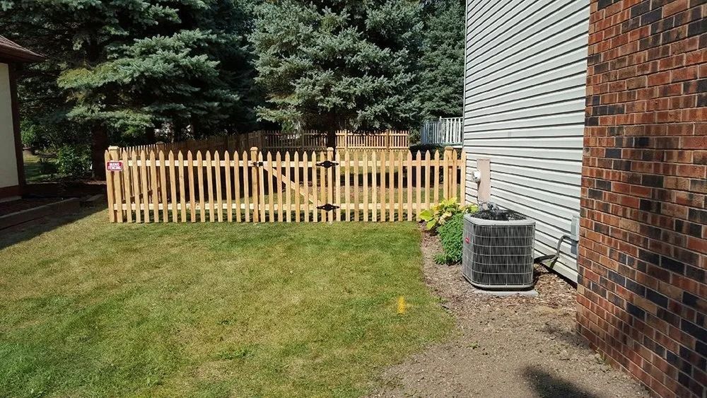 A backyard lawn with a wooden picket fence, gate, and central air conditioning unit against the side of a house.