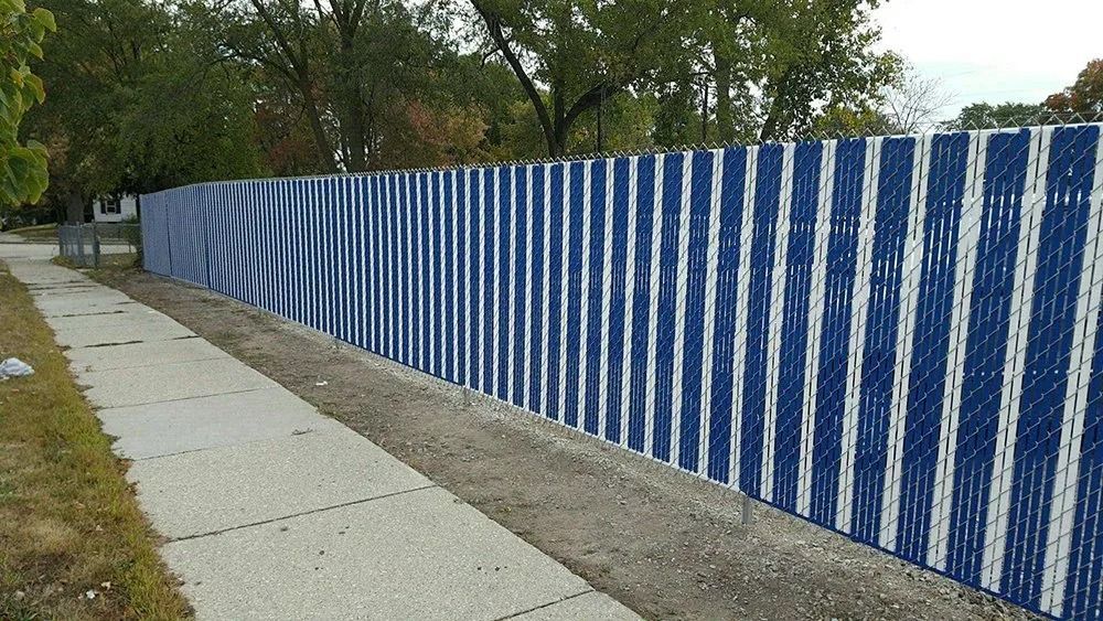 A sidewalk runs along a chain-link fence covered in vertical blue and white privacy slats, with trees in the background.