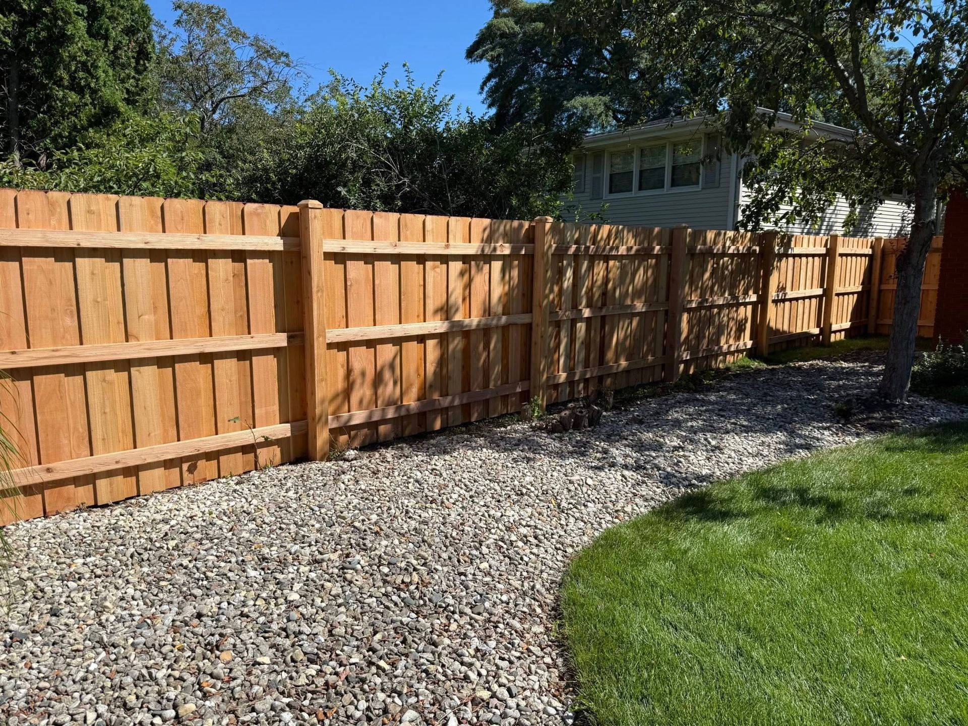 A new, tall, light-brown wooden fence stands along a gravel path next to a green lawn on a sunny day.