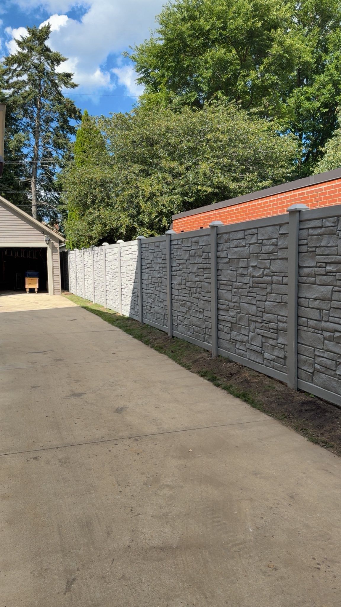 A concrete driveway leads to a garage beside a tall, grey faux-stone fence under a blue sky with green trees.