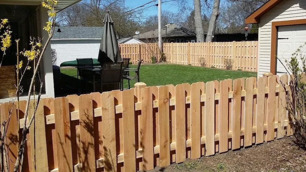 A light-colored wooden picket fence borders a backyard with a patio set and a green lawn on a sunny day.