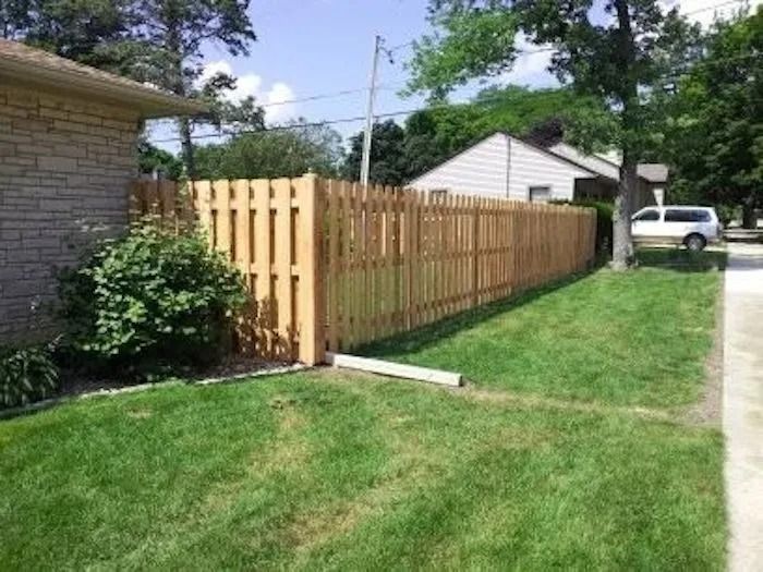 A new light-colored wooden picket fence stands between a brick house and a grassy yard on a sunny day.