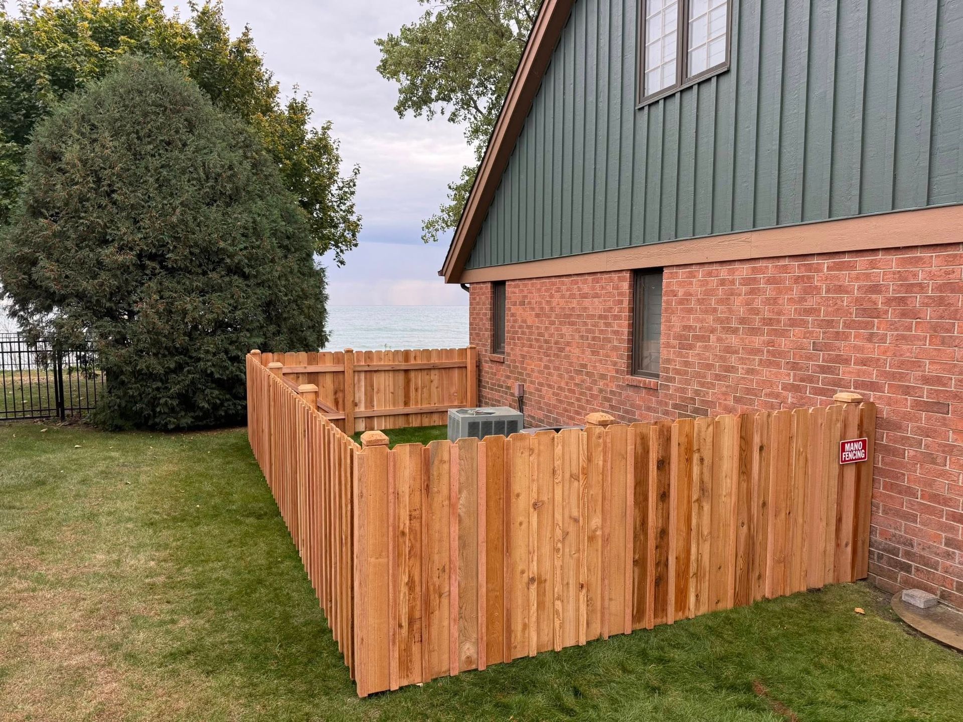 A cedar privacy fence encloses an air conditioning unit against the brick side of a house with green siding.