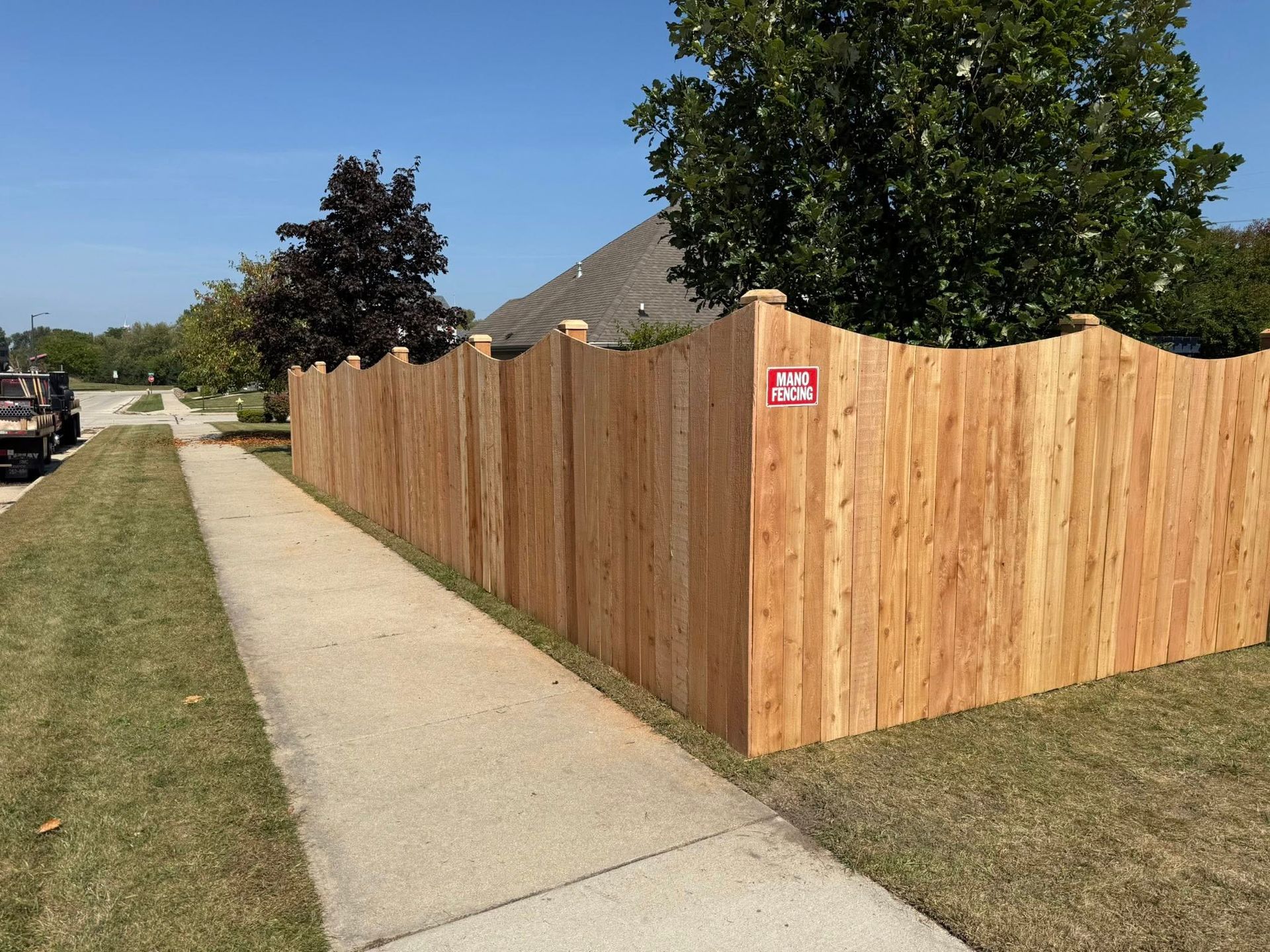 A newly installed, scalloped-top cedar wood privacy fence runs along a sidewalk bordering a residential yard.