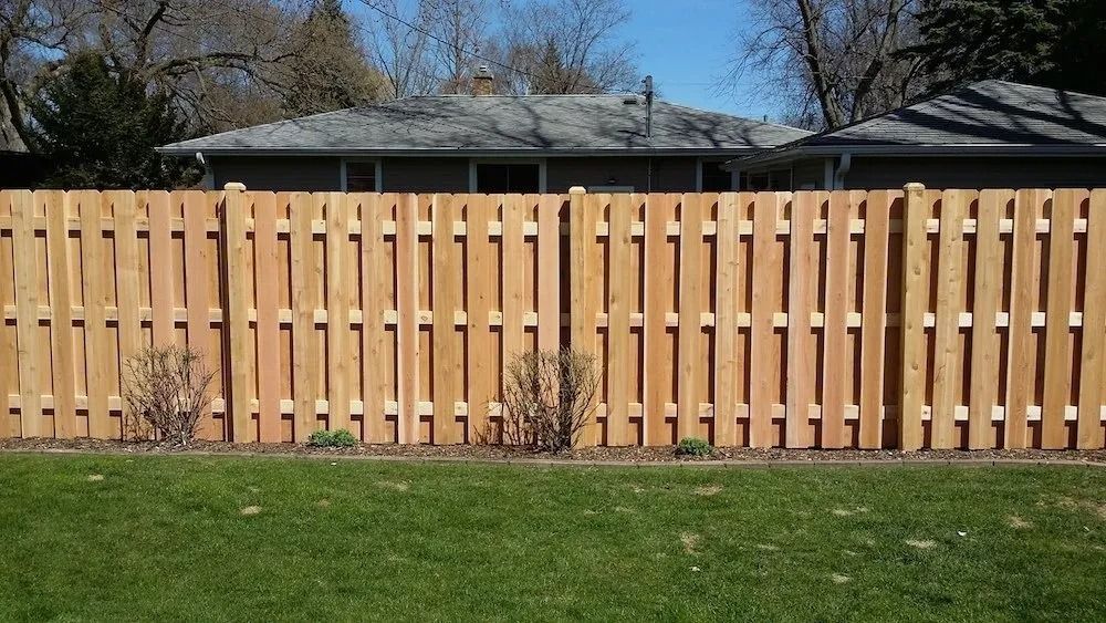 A wood fence with a natural finish separates a lush green lawn from the blurred silhouette of a house and trees.