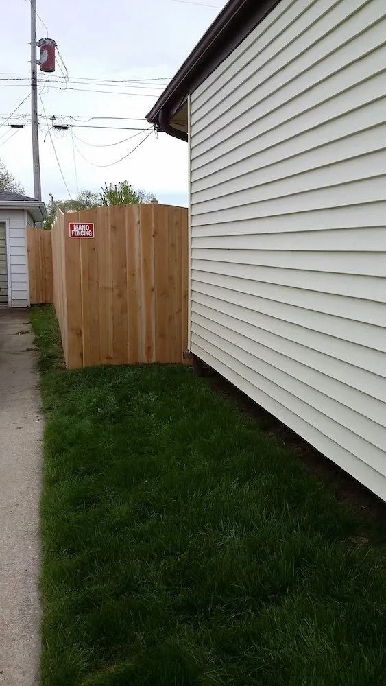 A side view of a house with beige siding next to a new wooden privacy fence and a grassy strip bordering a concrete path.