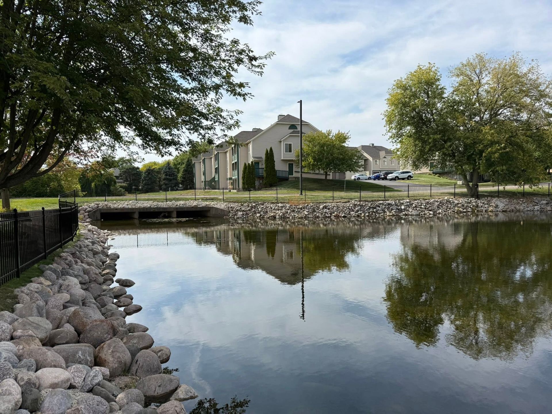 A calm pond with a rock-lined bank reflects a residential building and surrounding trees under a blue sky.