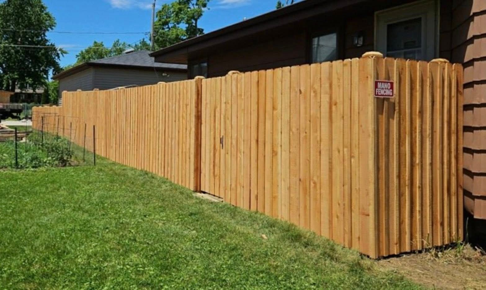 A tall wooden privacy fence runs along the side of a brown house, bordering a green lawn on a sunny day.