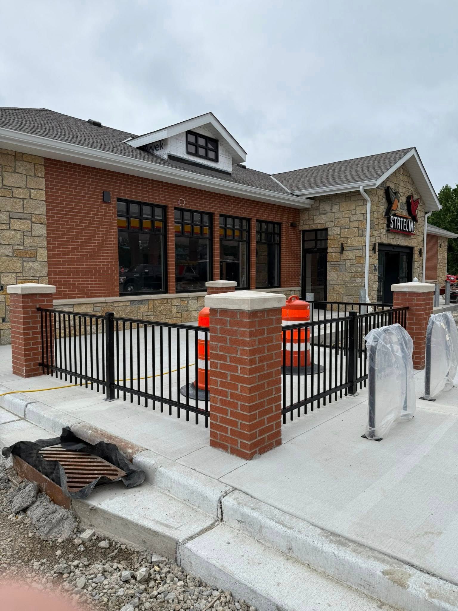 A newly constructed brick and stone building with an outdoor patio enclosed by black metal fencing under a cloudy sky.