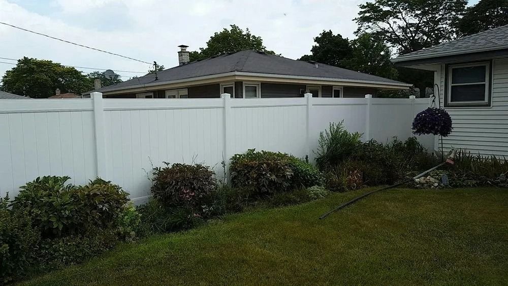 A white vinyl privacy fence runs behind a manicured lawn with shrubs and a house corner in a residential backyard.