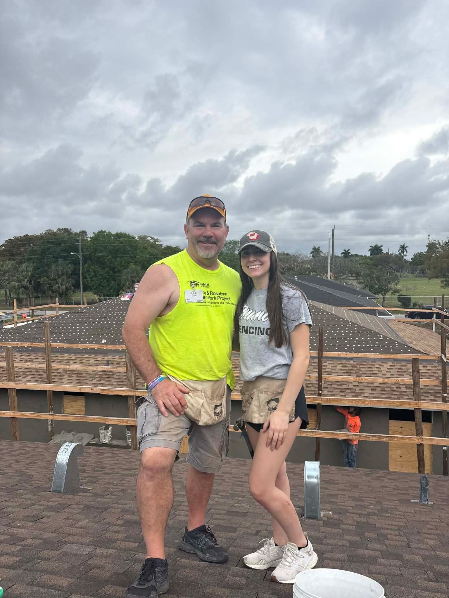 Two people in caps and work gear stand together on a rooftop construction site under a cloudy sky.