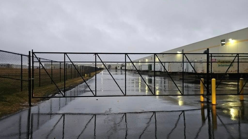 A black chain-link gate blocks a wet, paved driveway leading to a row of commercial warehouse loading docks.