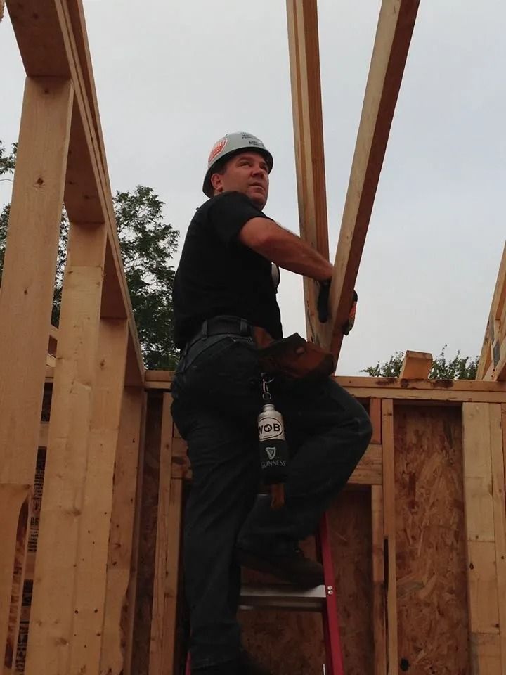 A person in a hard hat and tool belt stands on a red ladder, reaching up to support a wooden beam in a building frame.