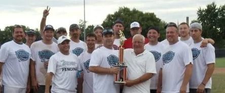 A baseball team in matching white shirts posing for a photo while holding a championship trophy on a field.