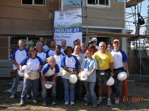 A large, diverse group of volunteers in matching t-shirts poses for a photo in front of a house under construction.