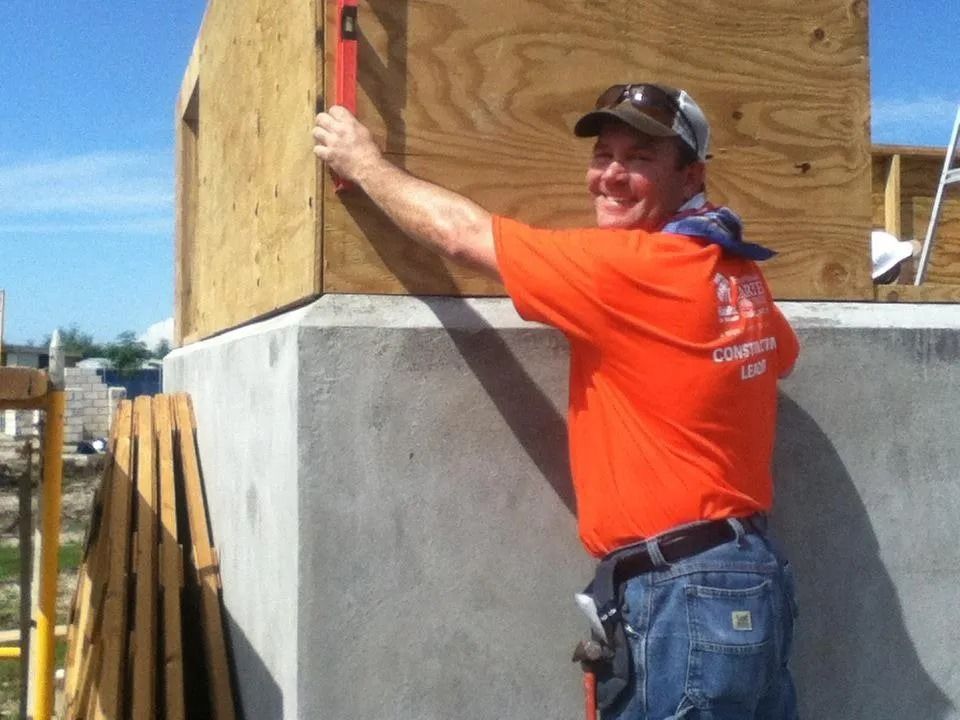 A person in an orange shirt smiles while using a level to check the alignment of plywood on a concrete building foundation.
