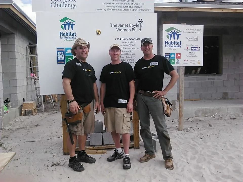 Three people in matching black shirts and work attire stand in front of a Habitat for Humanity construction site sign.