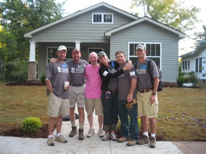 Six people in casual attire stand in front of a newly built, light-gray house with white trim.