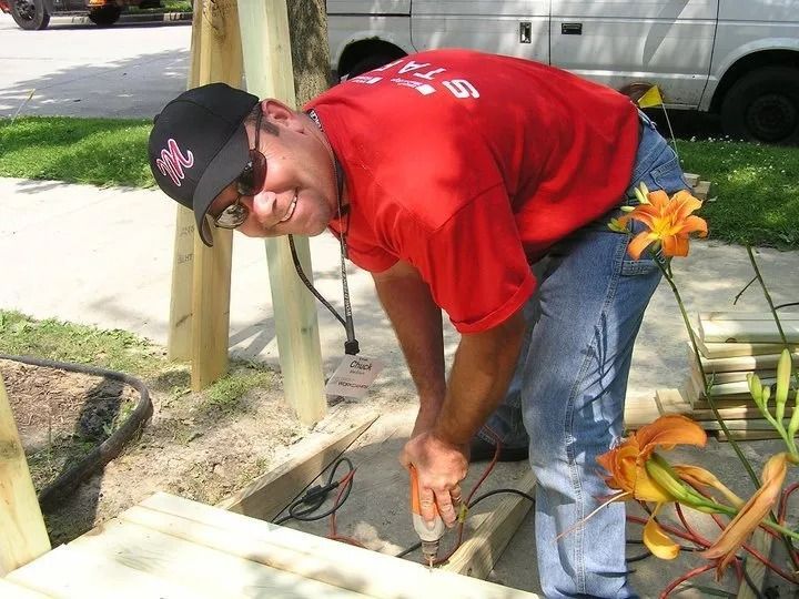 A person in a red shirt and black cap smiles while using a power drill on a wooden deck structure outdoors.