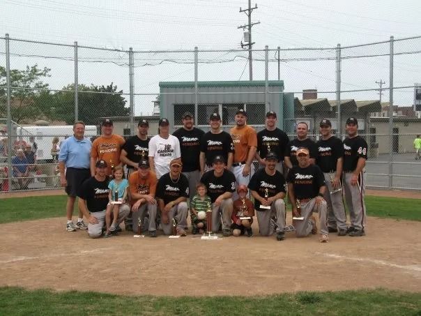 A baseball team in matching black shirts poses for a group photo on a dirt field in front of a chain-link fence.
