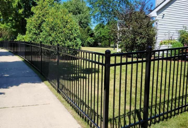 Black metal fence along a sidewalk beside a grassy yard and house on a sunny day