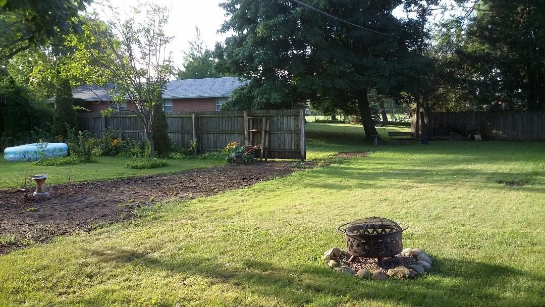 A backyard with a fire pit surrounded by rocks, a long dirt patch, a wooden fence, trees, and a house in the background.