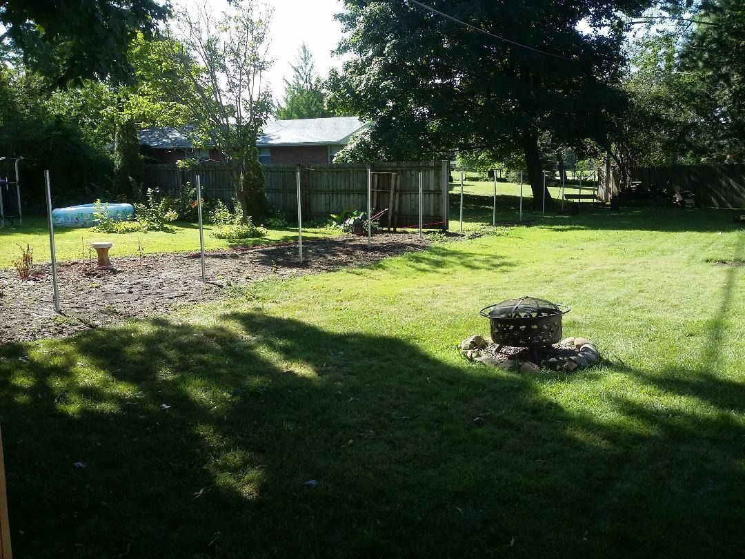 A backyard with a large, dark-mulched garden bed, a fire pit in the grass, and a shed in the background under large trees.