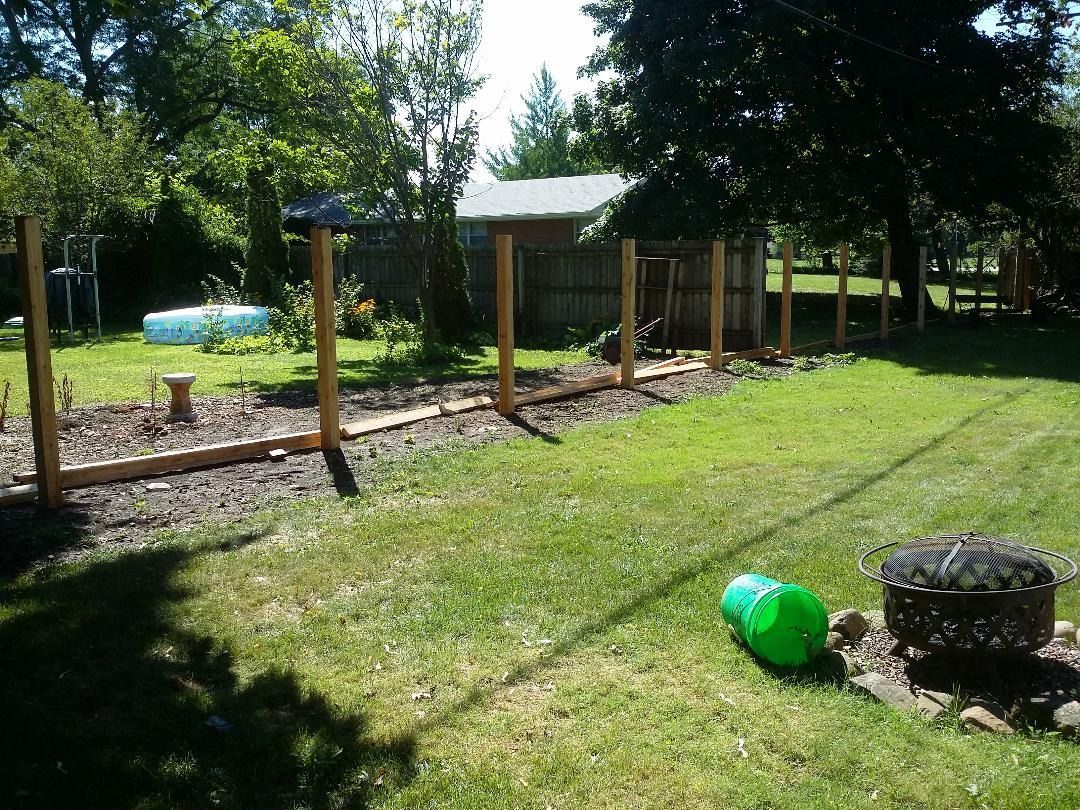 A row of wooden fence posts stands in a sunny backyard, with a garden bed in the foreground and a fire pit and green bucket.