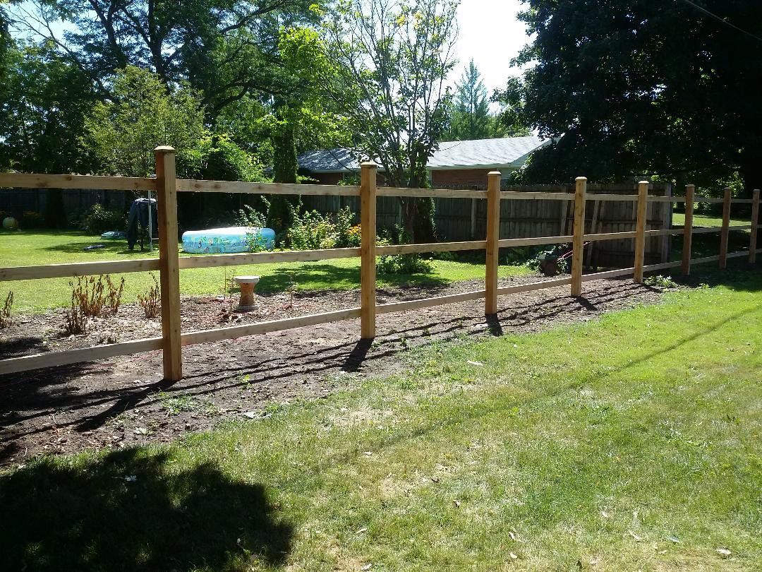 A three-rail wooden fence lines a garden bed with mulch, set against a green lawn and trees on a sunny day.