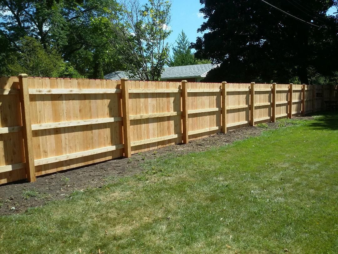 A new, light-brown wooden privacy fence stands between a mowed green lawn and dense trees under a clear blue sky.