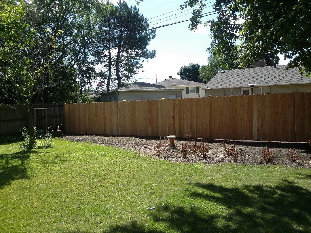A backyard scene featuring a new wooden fence separating a manicured lawn from a flower bed with sparse plants.