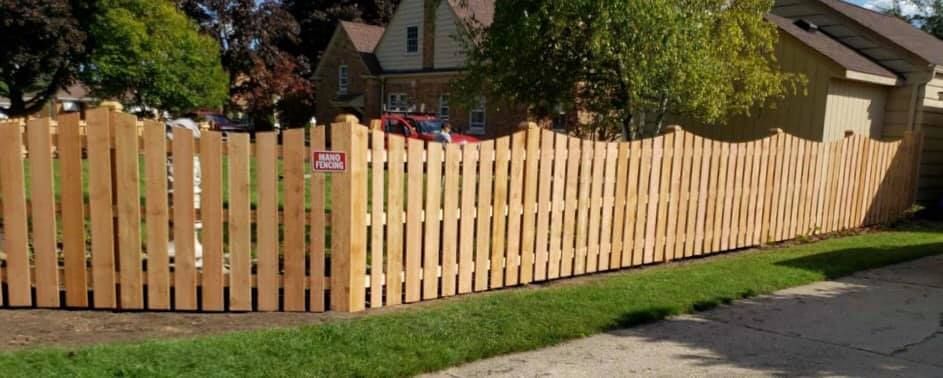 A newly installed wooden picket fence with a scalloped top edge sits in a residential yard beside a sidewalk.