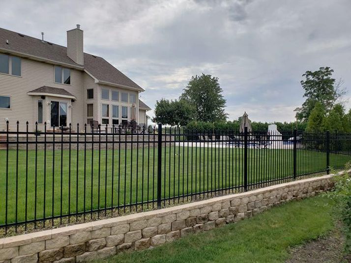 Large beige house with black iron fence and stone retaining wall beside a green lawn under cloudy sky