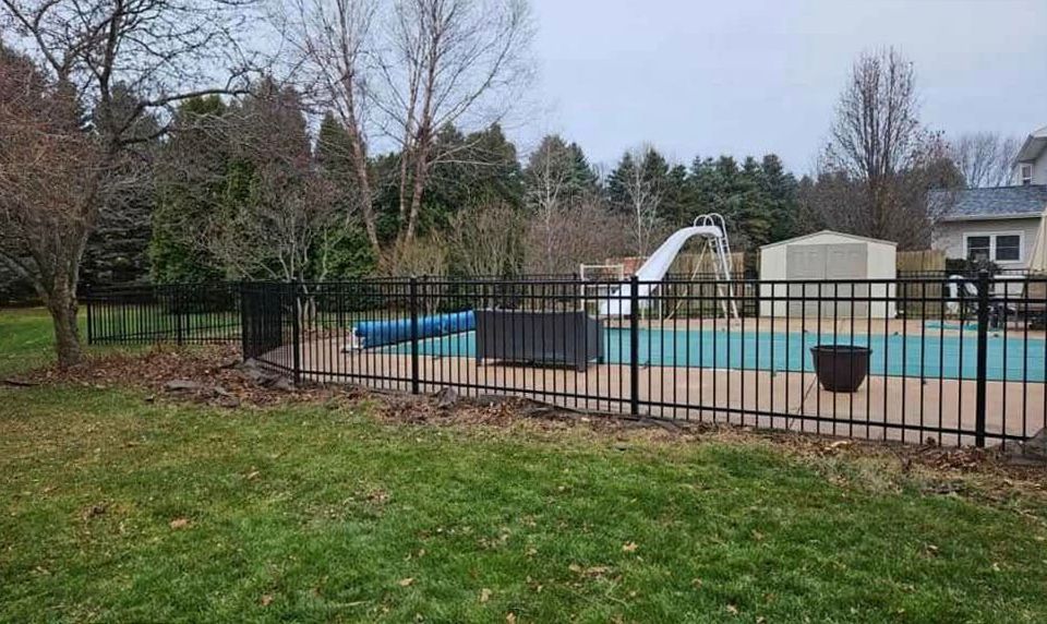 Backyard swimming pool behind black metal fence, with slide and pool cover in a wooded yard