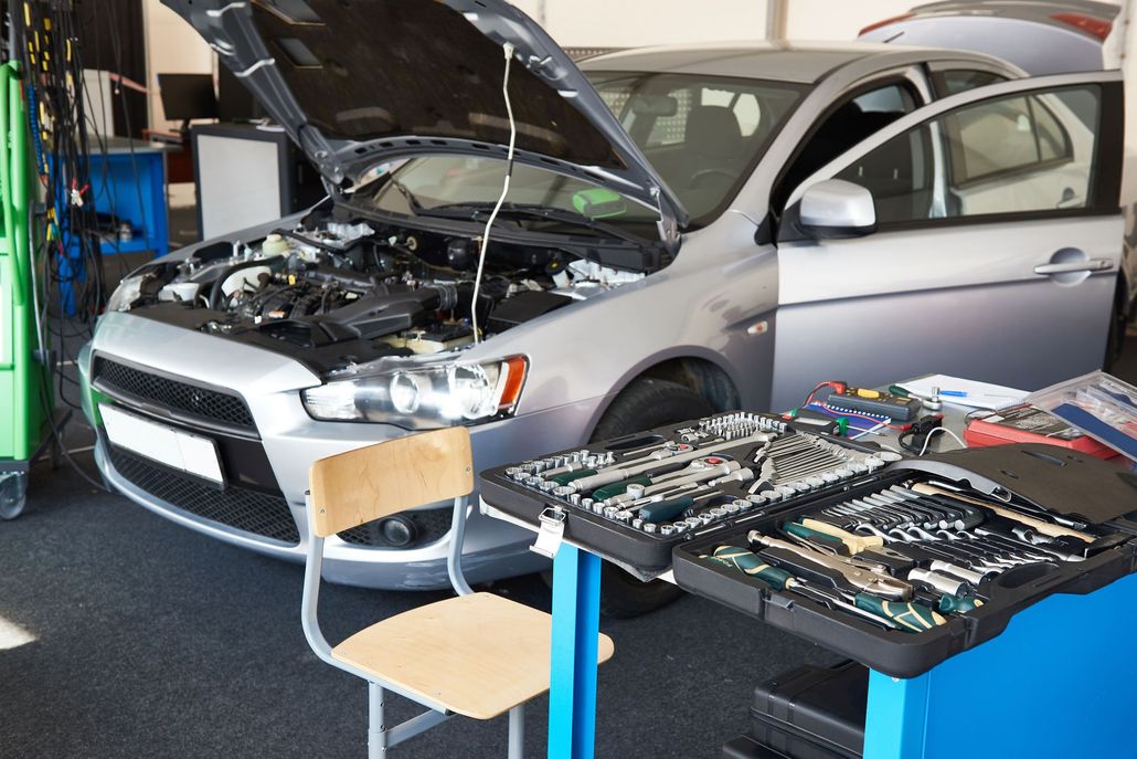 Silver car with hood open in a garage, with tool cases and a chair in the foreground