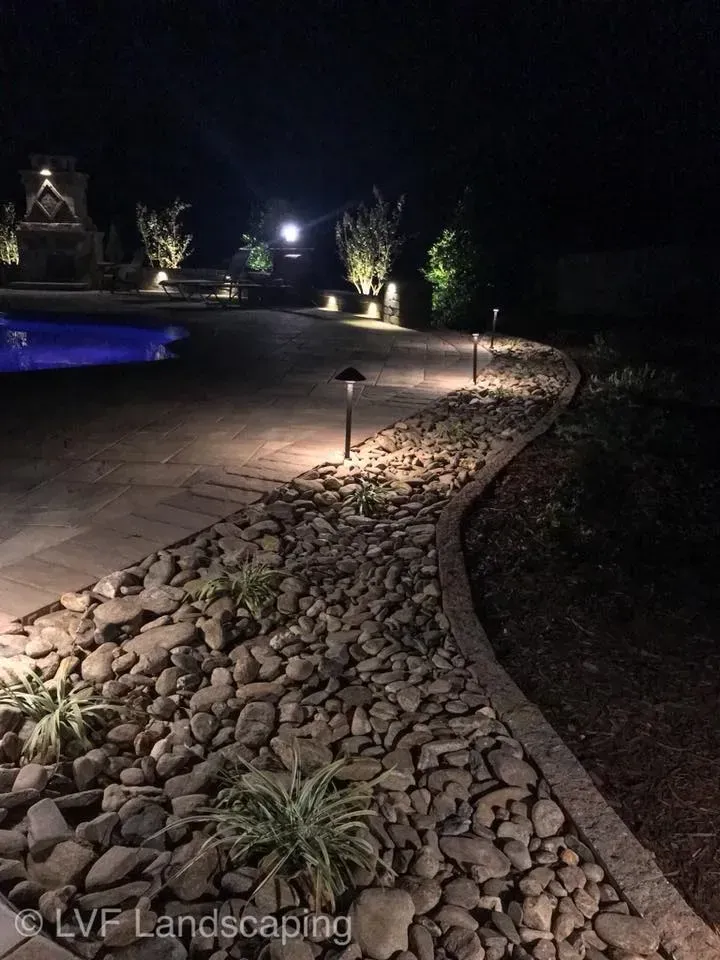 Pathway lined with rocks illuminated by landscape lighting at night, leading towards a pool and other illuminated features.