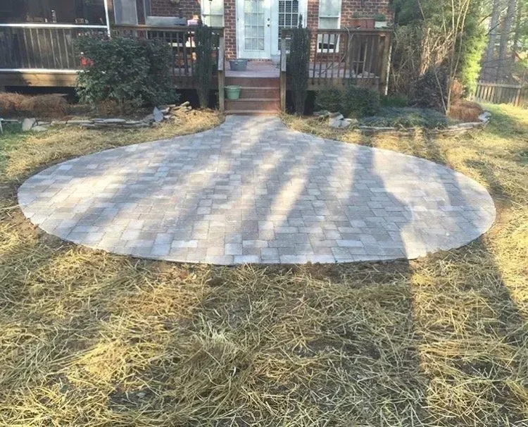 A circular brick patio in a yard, leading to a back deck. The grass surrounding the patio is brown.