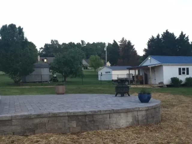 Brick patio overlooking a yard with sheds, a small house, and trees under a cloudy sky. A fire pit and potted plants are on the patio.