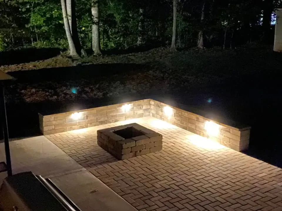 Brick patio at night with a square fire pit. Beige retaining wall has built-in lights, illuminating the area. Dark trees are in the background.