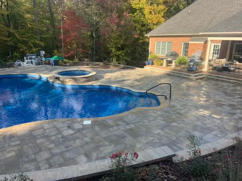 A backyard with a pool and spa, surrounded by a paved patio.  A brick house and fall foliage are in the background.
