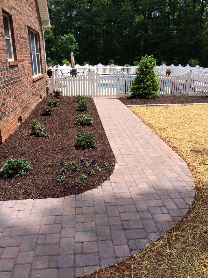 Brick pathway curves around a flower bed next to a brick house. A white picket fence and pool are visible in the background.