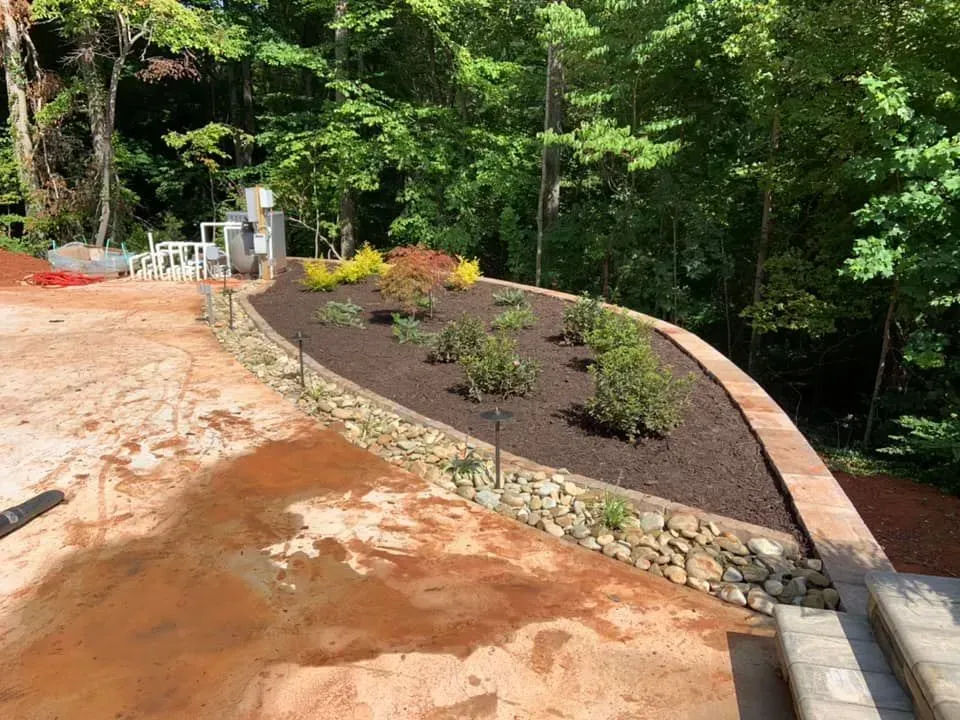 A raised garden bed filled with mulch and plants, surrounded by a stone wall, next to a patio and a wooded area.
