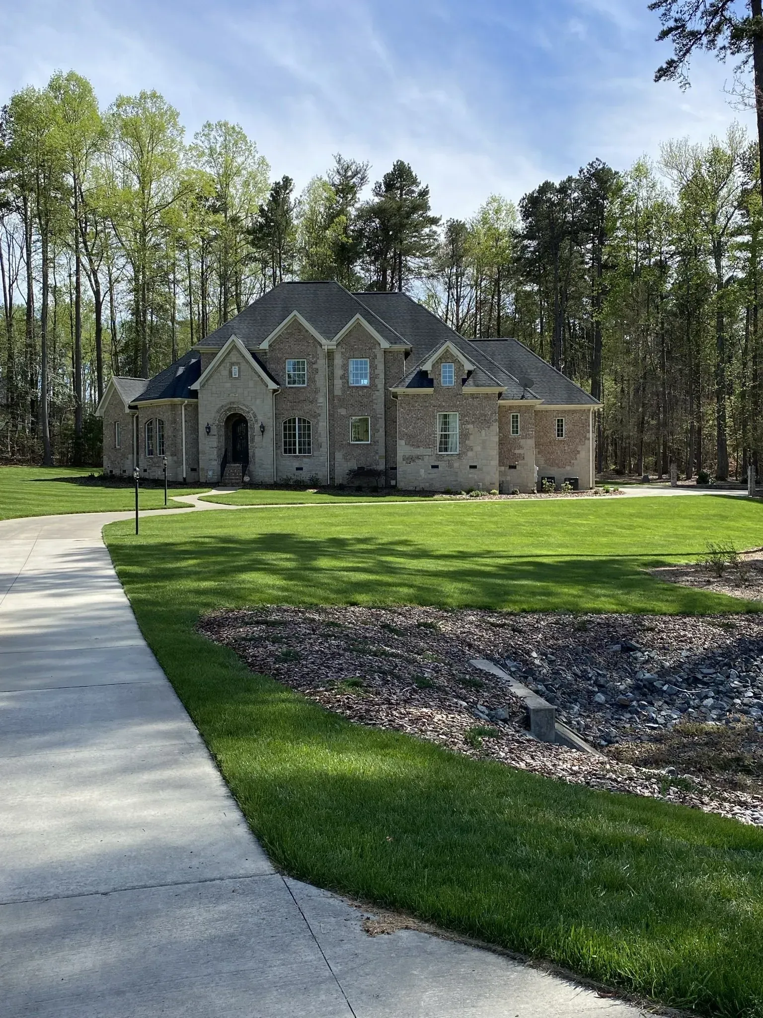 Large two-story brick house surrounded by a green lawn and tall trees. A paved walkway leads towards the entrance.