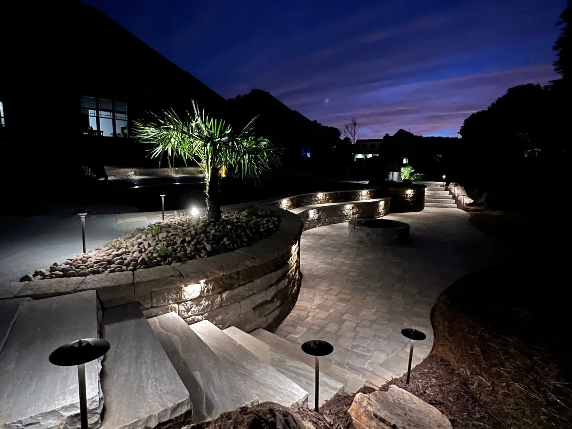Night view of a tiered outdoor patio with stone steps, pathways, and subtle lighting illuminating a palm tree and surrounding landscape under a twilight sky.