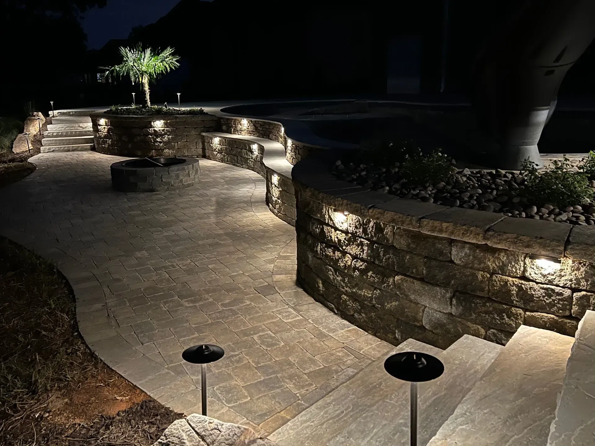 Dark pathway and stone walls illuminated by lights, with a tree and steps in a nighttime setting.