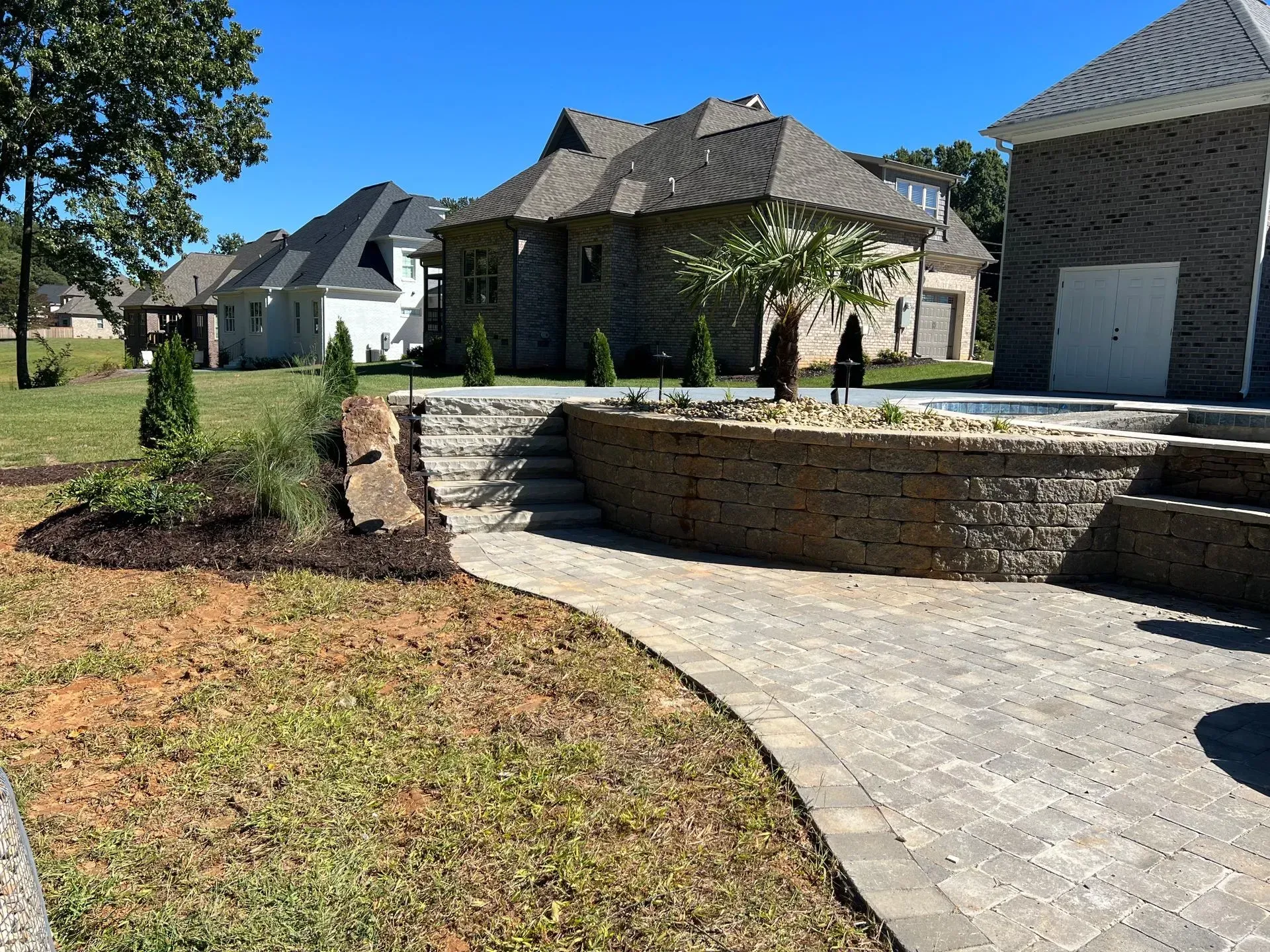 A landscaped yard with stone retaining walls, steps, and a paved pathway leading to a house under a blue sky.