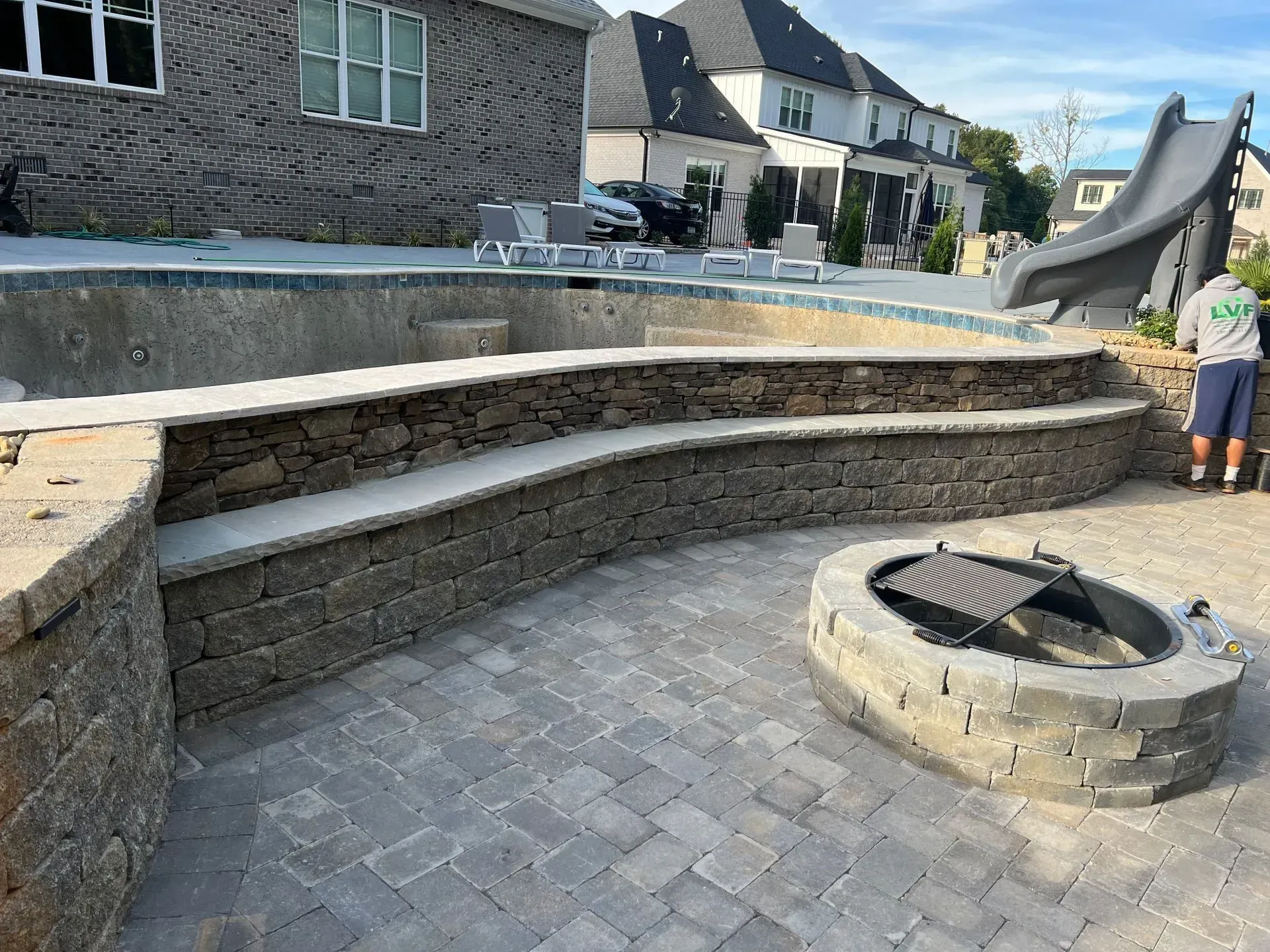 A backyard with a pool, seating wall, fire pit, and slide. A person stands near the slide, and the patio is made of pavers.