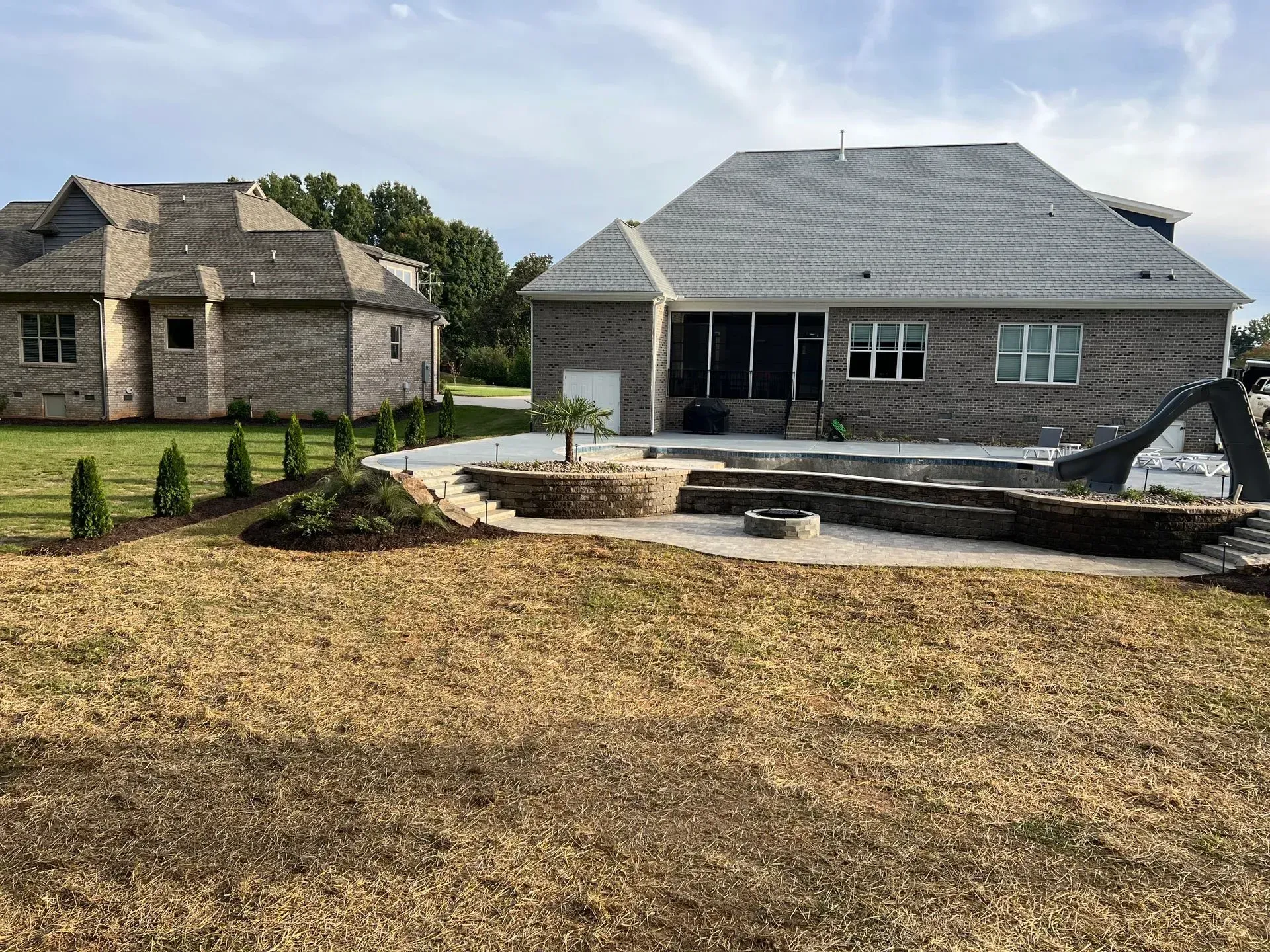 Backyard with a pool, patio, and landscaping, with a brick house and outbuilding. Dry, brown grass covers much of the yard.