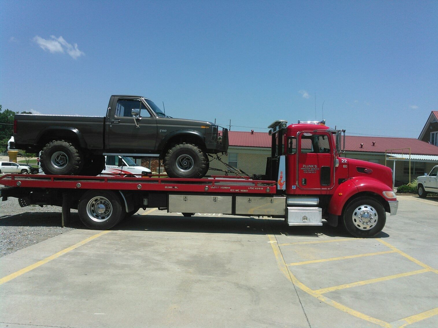 Red tow truck carrying a dark green pickup truck on a flatbed in a parking lot.