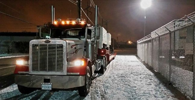 Snowy night scene with a semi truck parked beside a chain-link fence under bright lights